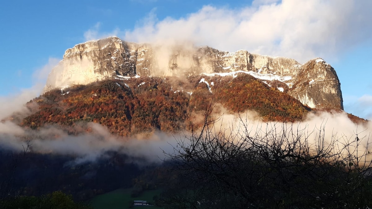 A majestic mountain is portrayed, with its rugged cliffs partially shrouded in mist. The autumn foliage adds warm tones to the lower landscape, while snow caps the summit, contrasting against the vibrant colors of the trees below. The sky is painted in soft hues of blue and white.