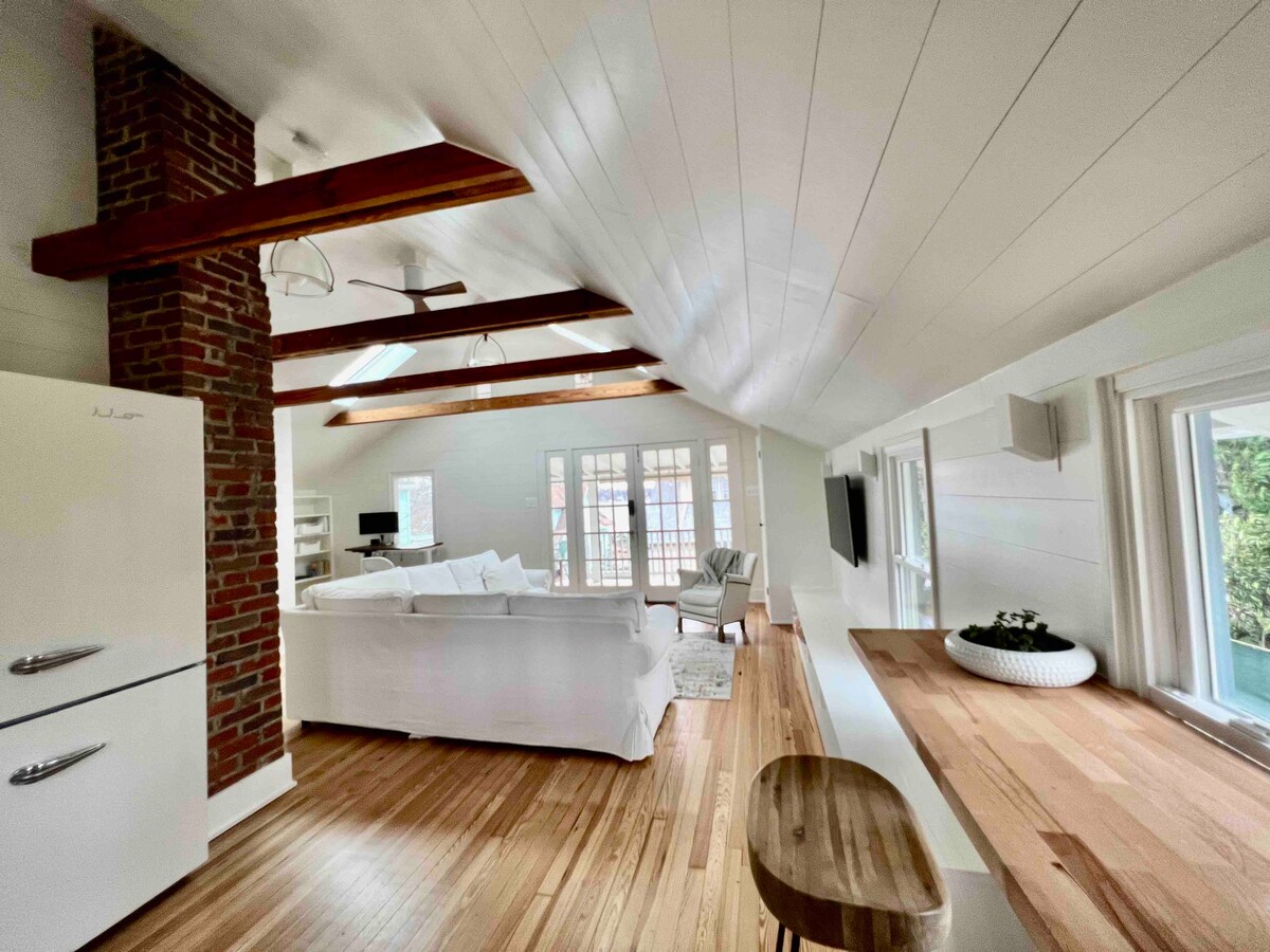 The airy living space features high ceilings with exposed wooden beams. A comfortable white sofa is positioned near large windows, allowing natural light to fill the room. A wooden table provides a casual dining option, while a vintage-style refrigerator adds character to the kitchen area.