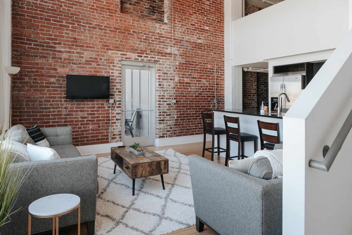 The living area features unique brick walls with high ceilings, creating an open feel. Two gray sofas are positioned around a wooden coffee table, set on a soft rug. A small round table is visible, and a sleek kitchenette is located in the background, alongside a flat-screen television.