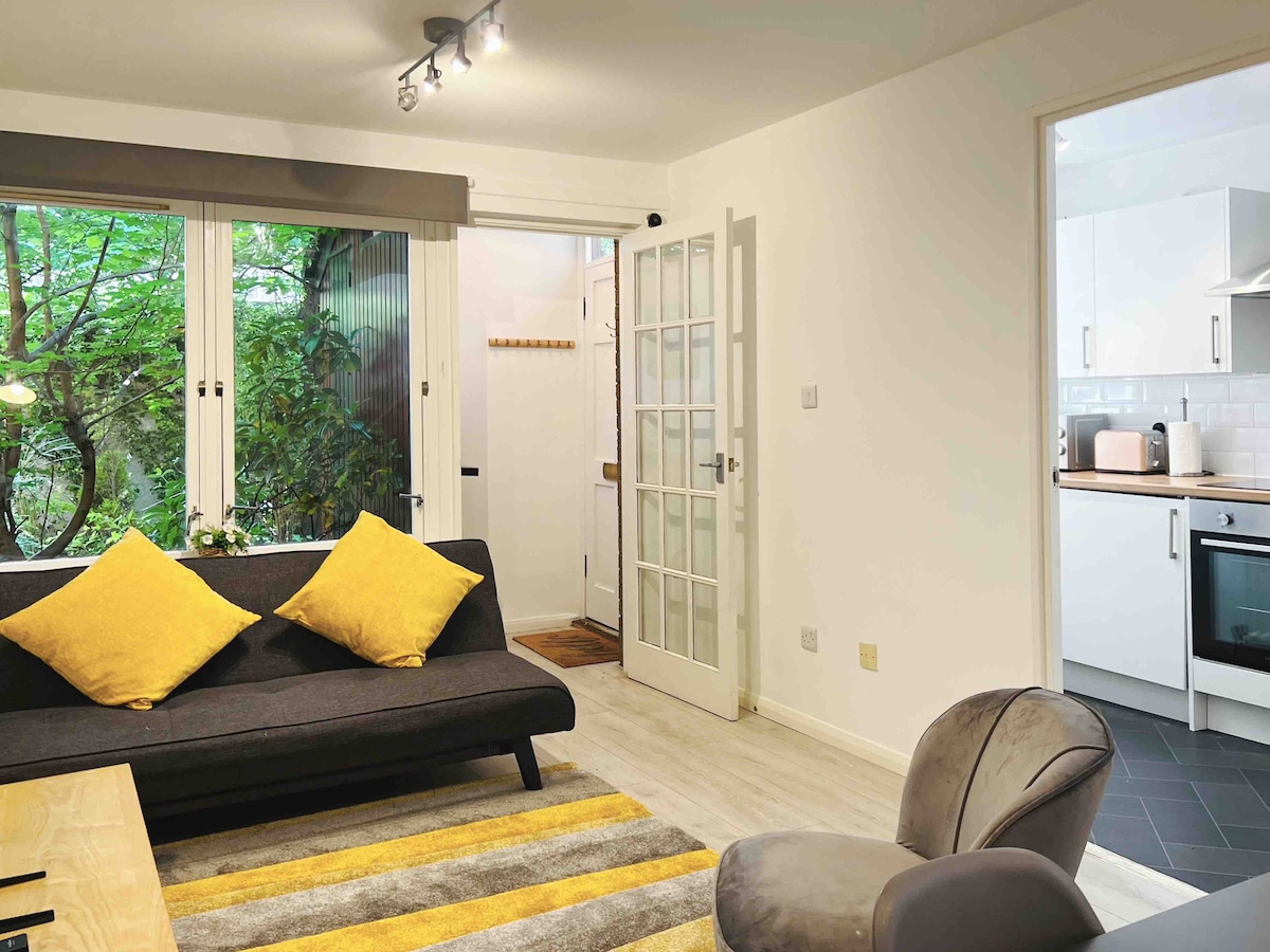 The living area features a modern sofa adorned with yellow cushions, positioned on a patterned rug. Natural light enters through glass doors, revealing greenery outside. A compact dining area is visible, leading to a well-equipped kitchen with white cabinetry and modern appliances.