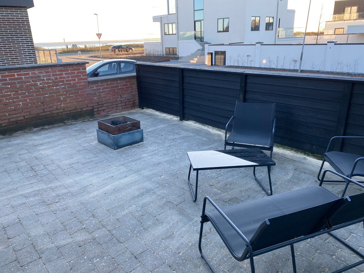 A private terrace features a fire pit surrounded by two black chairs and a small triangular table. The space offers views of the surrounding area, with modern buildings visible in the background.