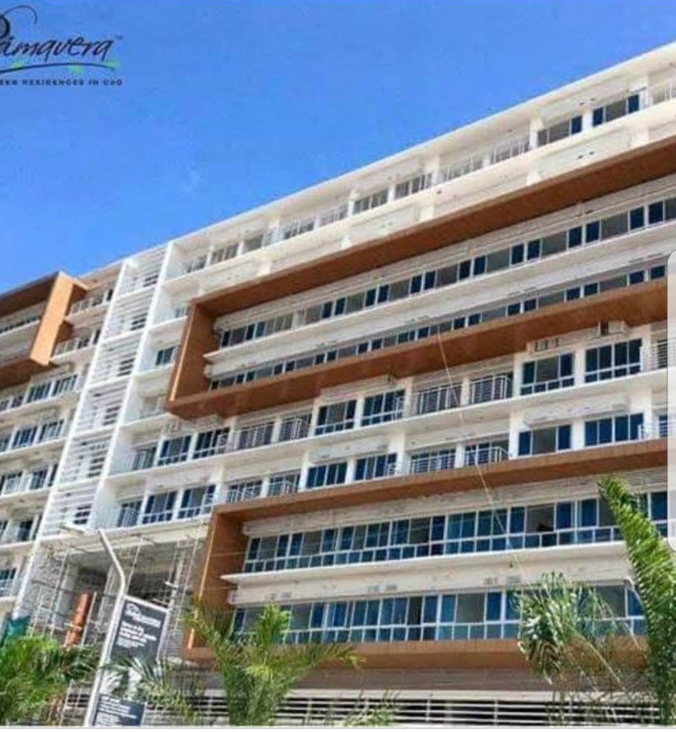 A modern condominium building features a blend of white and warm-toned facades with multiple floors of large glass windows. Lush greenery is visible in the foreground, creating a refreshing contrast to the architectural lines.