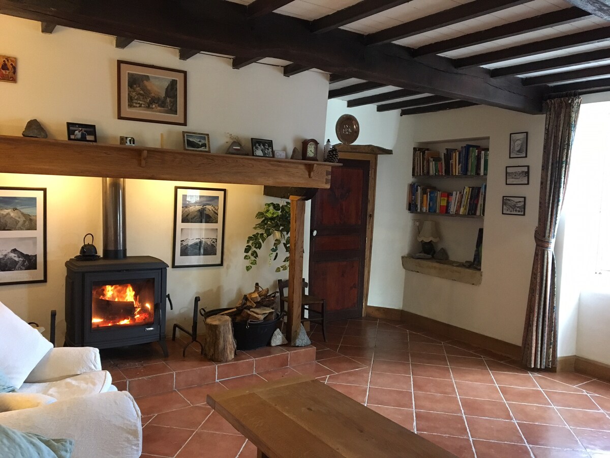 A cozy sitting area features a wood-burning stove set against a backdrop of framed photographs. Natural light fills the room through a window adorned with curtains. Bookshelves with an eclectic selection of books are mounted on the wall, enhancing the homey ambiance.