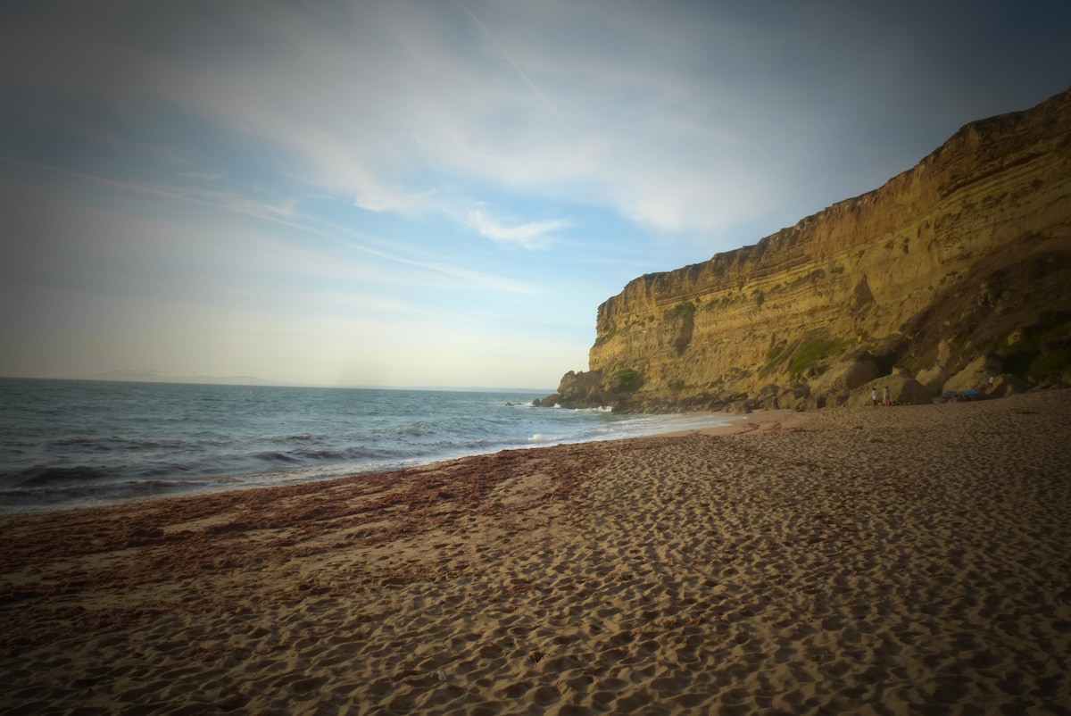 A sandy beach stretches along the coastline, with gentle waves lapping at the shore. A cliff rises in the background, showcasing layered rock formations under a clear blue sky. The scene conveys a serene atmosphere, ideal for relaxing by the sea.