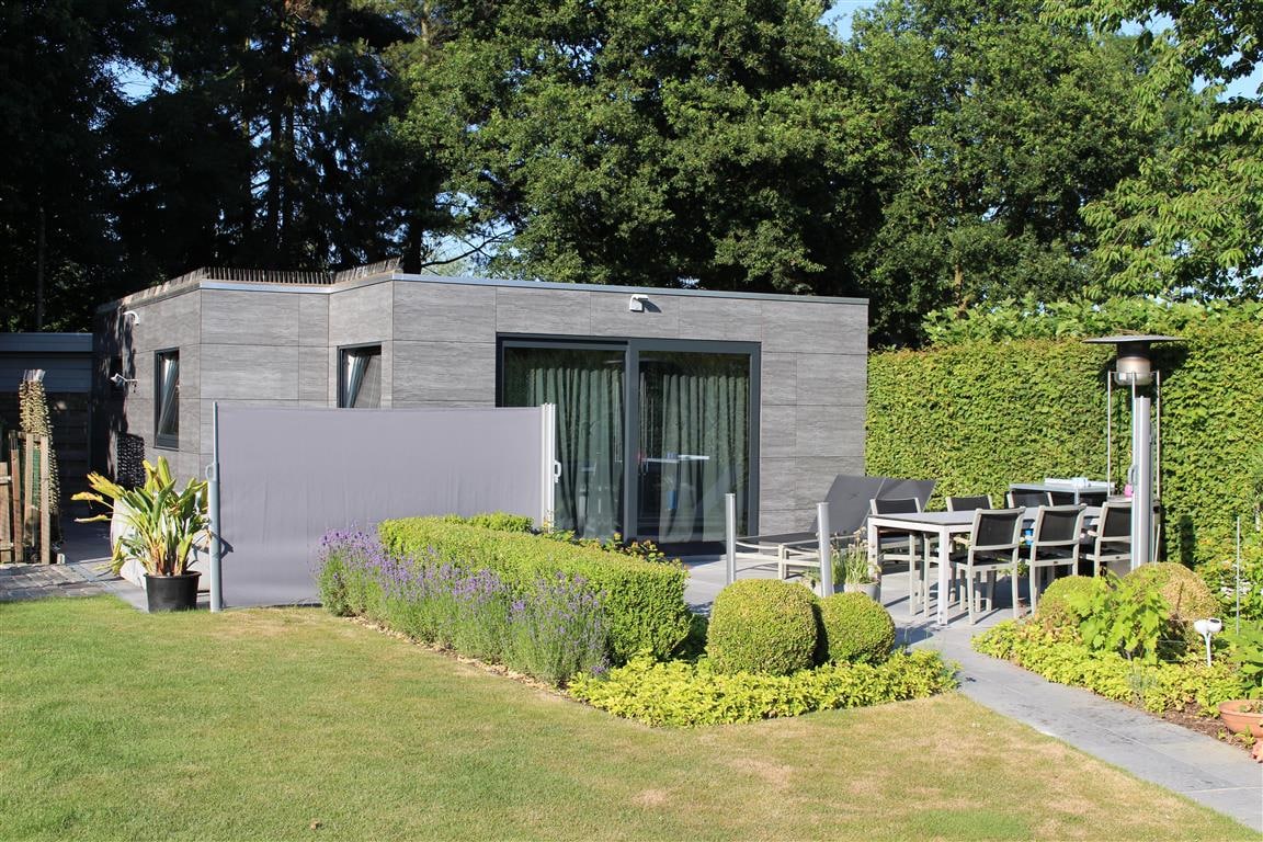 A modern detached guesthouse is set within a lush garden, surrounded by neatly trimmed hedges. An outdoor seating area is visible, featuring a table and chairs. A pathway made of stones leads toward the entrance, with patches of greenery and flowers adding charm.