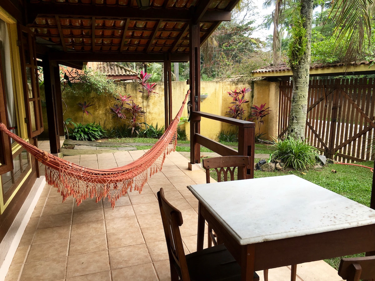 A covered outdoor patio is featured, showcasing a light-colored table and wooden chairs. A vibrant hammock is hung for relaxation. Lush greenery and colorful plants are visible in the background, adding to the tranquil setting.