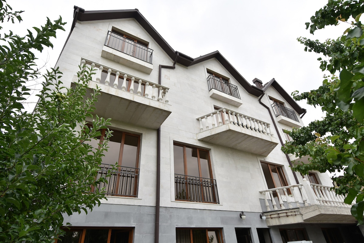 The three-story building features several large windows, each with stylish railings. Balconies extend from the second and third levels, providing outdoor space with views. The exterior is complemented by surrounding greenery, including trees that enhance the natural setting.