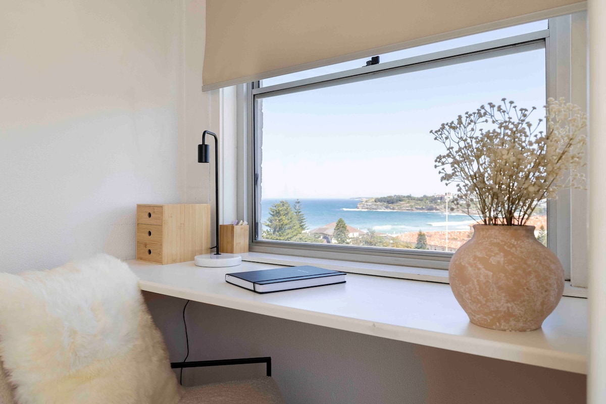 A serene workspace is seen by a window that offers an ocean view. A wooden desk holds a notebook and a simple lamp, while a decorative vase with dried flowers adds a touch of warmth. Soft textures enhance the inviting atmosphere.