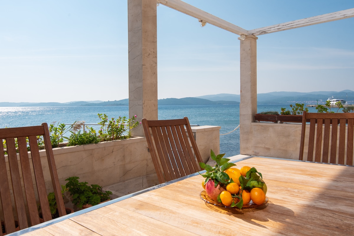 A wooden dining table is set on a terrace overlooking the Peljesac channel. A bowl of assorted fresh fruits rests centrally on the table. The view features calm waters and distant hills under a clear blue sky, framed by supportive columns and greenery.