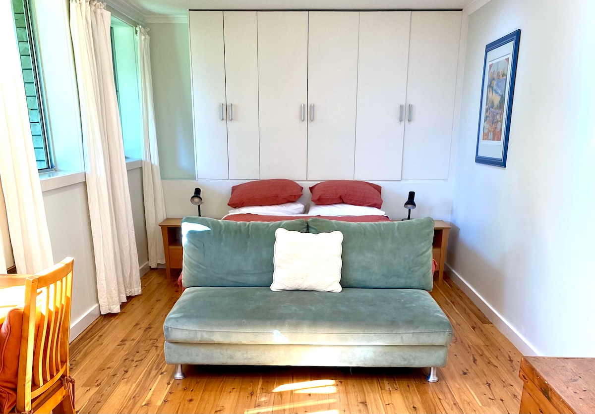 A cozy sleeping area features a spacious bed with soft bedding, flanked by two bedside tables and modern lamps. A green sofa is positioned in front, while light filters in through large windows with sheer white curtains, illuminating the warm wooden floor.