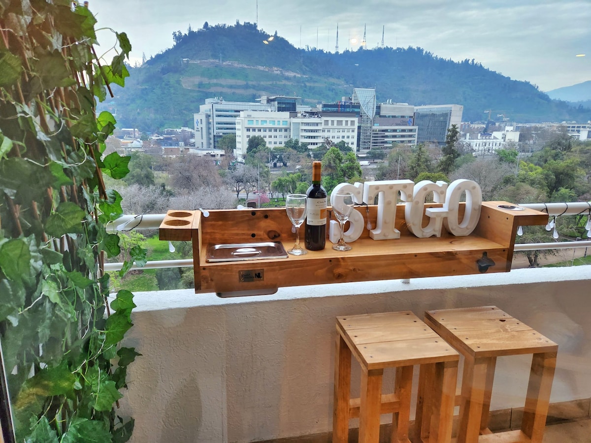 A balcony is presented, featuring a wooden bar with letters spelling 'STGO.' A bottle of wine and two glasses are arranged atop the bar. Two wooden stools provide seating. The backdrop includes a scenic view of the tree-lined area and distant hills.