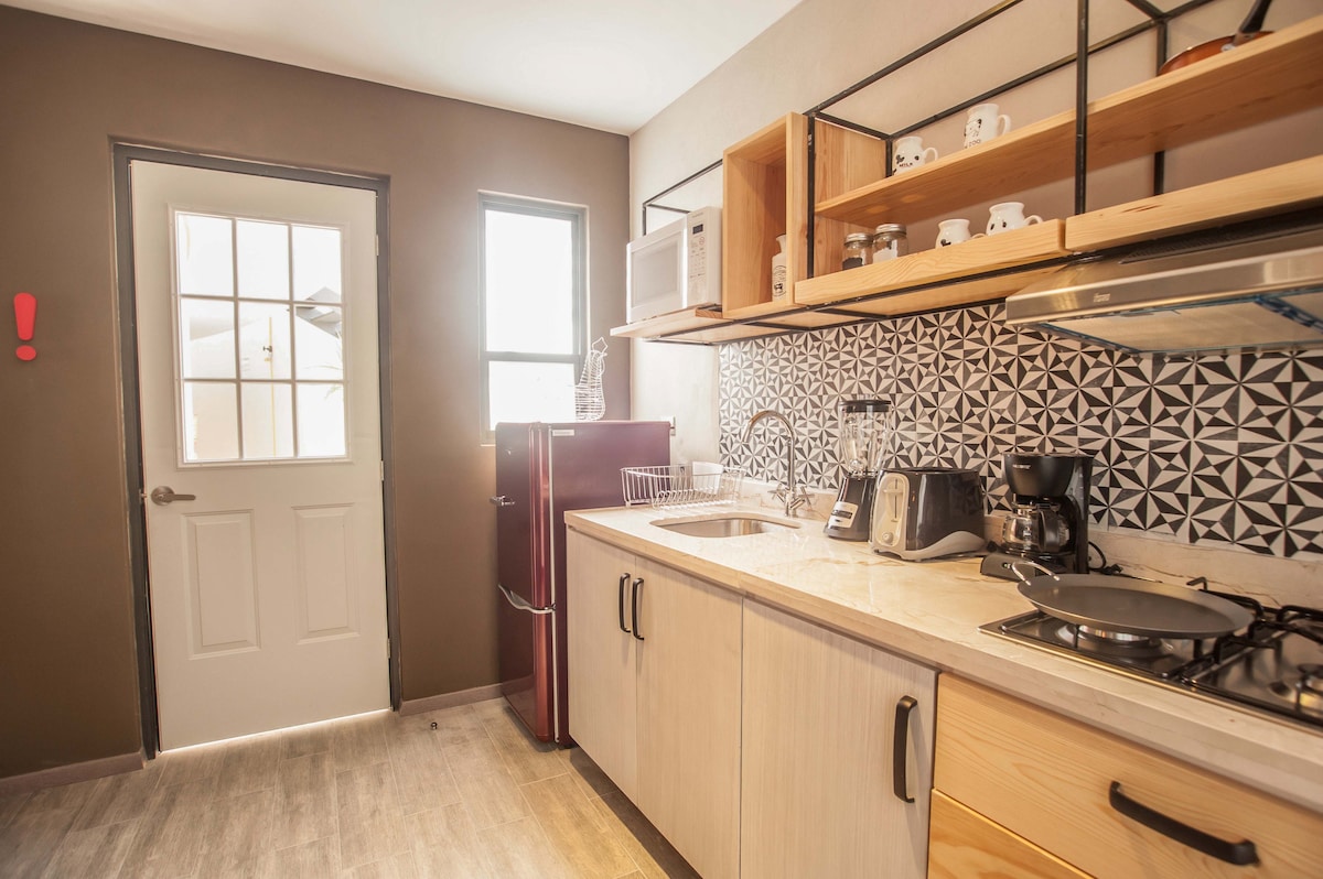 A modern kitchen features light wooden cabinetry and a marble countertop. Appliances include a red refrigerator, microwave, coffee maker, and stovetop. A patterned backsplash adds visual interest, while natural light floods in from a window above the sink, highlighting the kitchen's functional layout.