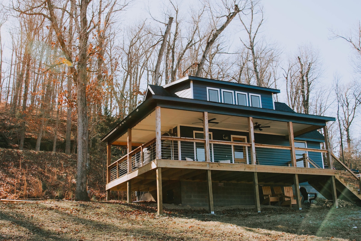 The cabin exterior is depicted, showcasing a modern design with a spacious front porch featuring wooden railings. The porch is supported by sturdy pillars and overlooks a natural landscape with tall trees. Sunlight filters through the branches, illuminating the scene.