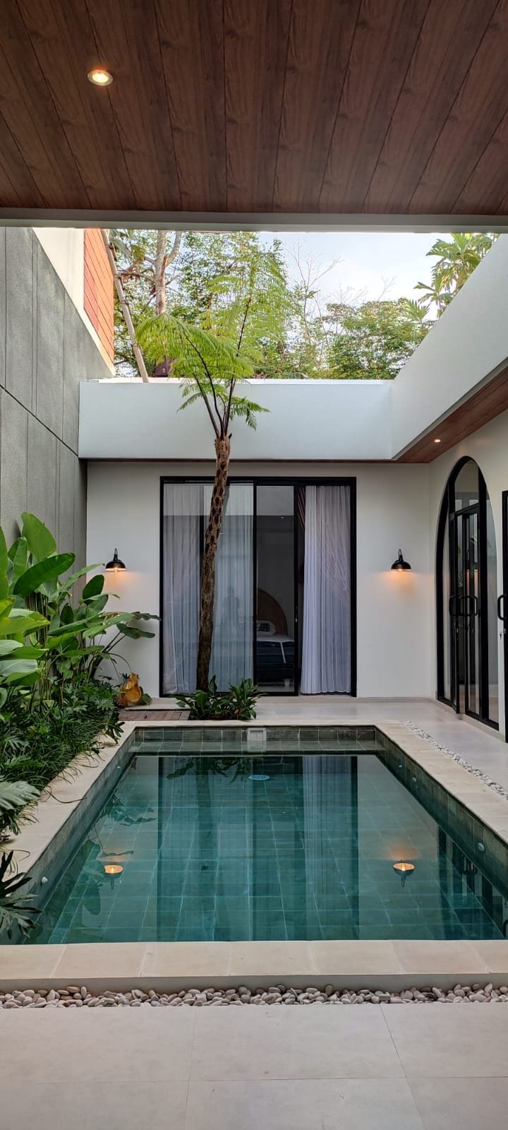 A serene courtyard is adorned with tropical plants surrounding a tranquil pool. Light reflects from the water's surface, while a slender tree adds height to the space. Large glass doors lead into the interior, offering glimpses of a cozy seating area.