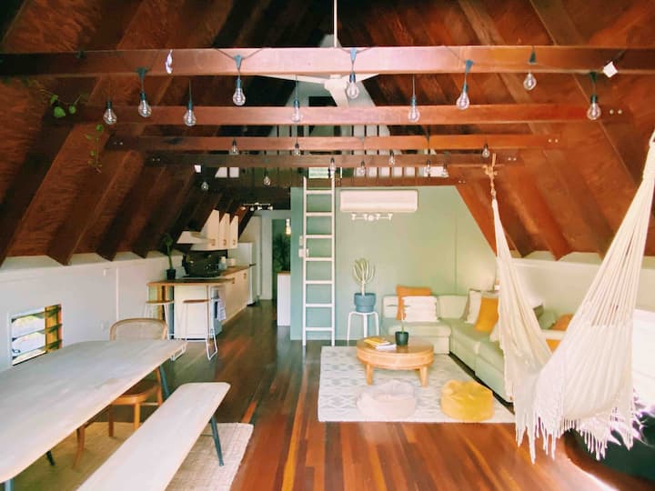 The image shows a coy, rustic loft space with slanted wooden ceilings. The interior features a mix of comfortable seating, including a hammock chair, and a dining table with benches. String lights hang from the beams, adding a warm, inviting atmosphere. The loft also has a small kitchenette, and a ladder leads up to an additional space. The decor is minimalist, with neutral tones and pops of yellow, creating a relaxed, airy vibe perfect for lounging and enjoying the space.