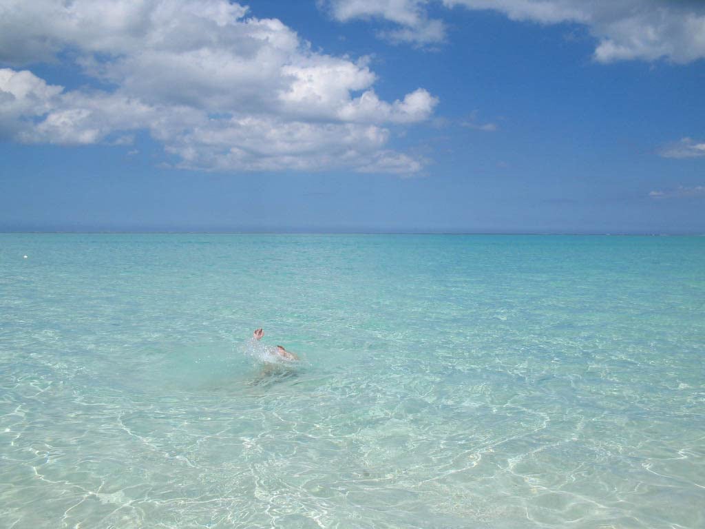 The clear, turquoise waters of the beach are depicted, where gentle waves lap against the shore. A swimmer is visible in the water, surrounded by the serene, shallow expanse of the ocean. The sky is dotted with soft clouds, contributing to the tranquil scene.