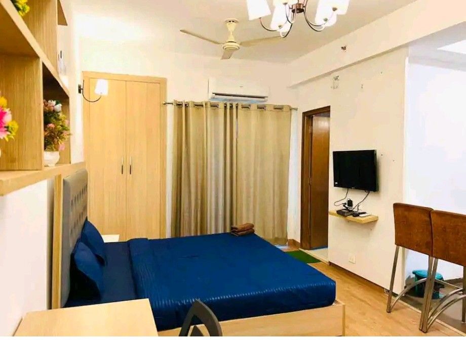 A neatly arranged sleeping area includes a spacious bed covered with a blue blanket. A television is mounted on the wall opposite the bed, while a pair of brown chairs and a small table are visible, enhancing the room's functionality.