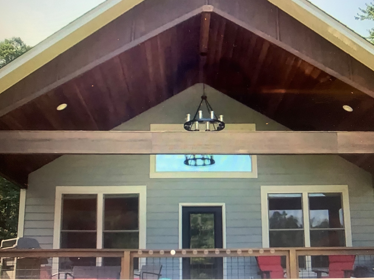 The exterior of a modern cabin is depicted, featuring a steeply pitched roof with wooden accents. A wide deck is visible, complemented by a decorative chandelier and several large windows, allowing ample natural light. The entrance is framed by two inviting chairs positioned on the porch.