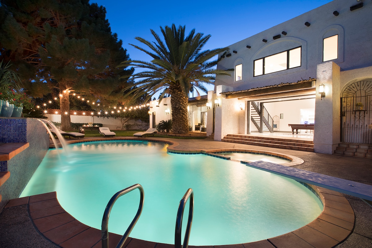 An inviting backyard features an illuminated pool with a gentle waterfall, surrounded by palm trees and landscaped greenery. Soft lighting from string lights creates a relaxed ambiance around the outdoor space. A spacious patio leads to the house, with a pool table visible nearby.