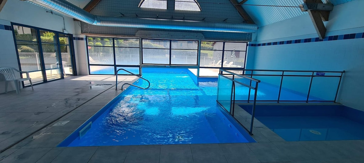 The indoor swimming pool area features clear blue water under a spacious roof with large windows, allowing natural light to fill the space. Various sections of the pool, including a shallow area and steps for easy access, are visible, creating an inviting atmosphere for relaxation.