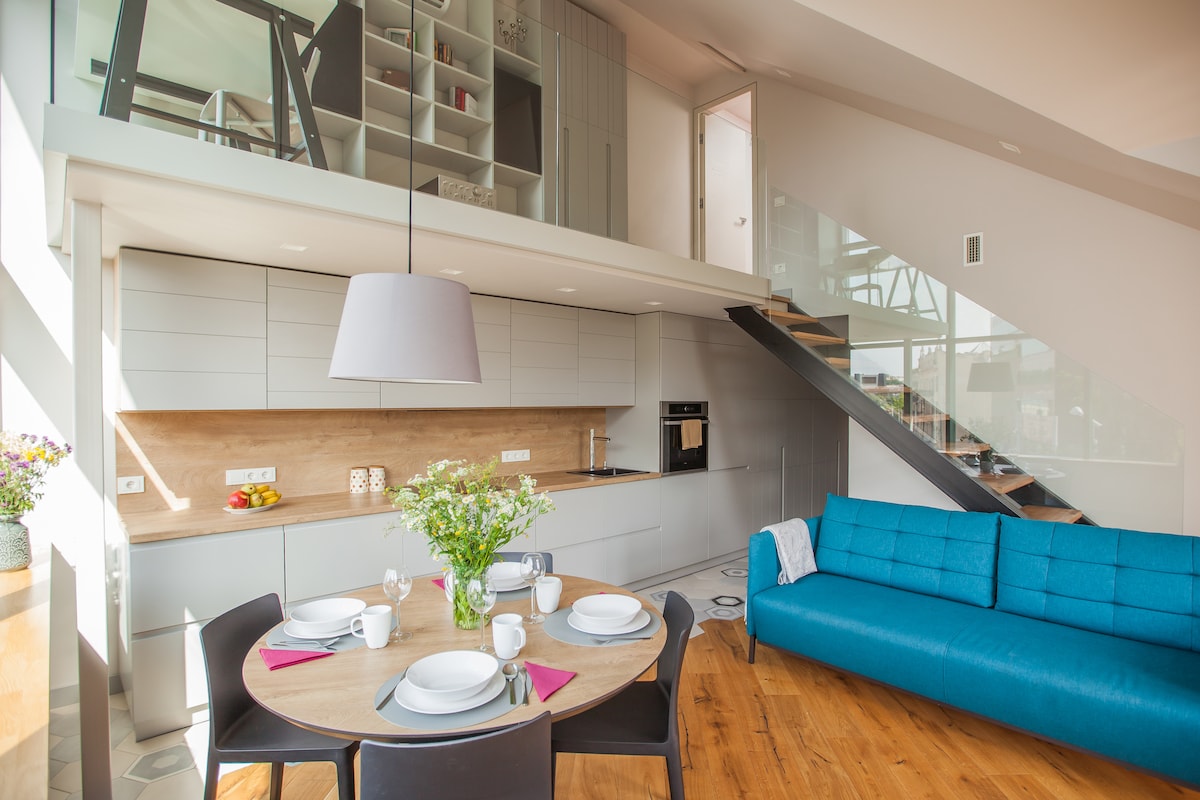 A modern open-plan living area features a round dining table set for four, with a vase of flowers as the centerpiece. The kitchen, equipped with sleek cabinetry, includes a built-in oven and countertop space. A turquoise couch adds a pop of color, while a staircase leads to an upper level.