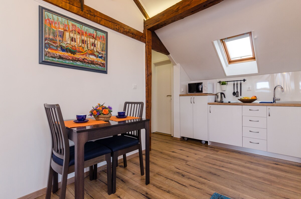 A dining area features a dark wooden table set for two, complemented by blue dishware. A kitchenette is visible in the background, equipped with a microwave and cooking utensils. Natural light enters through a skylight, enhancing the warm wood tones of the ceiling and flooring.
