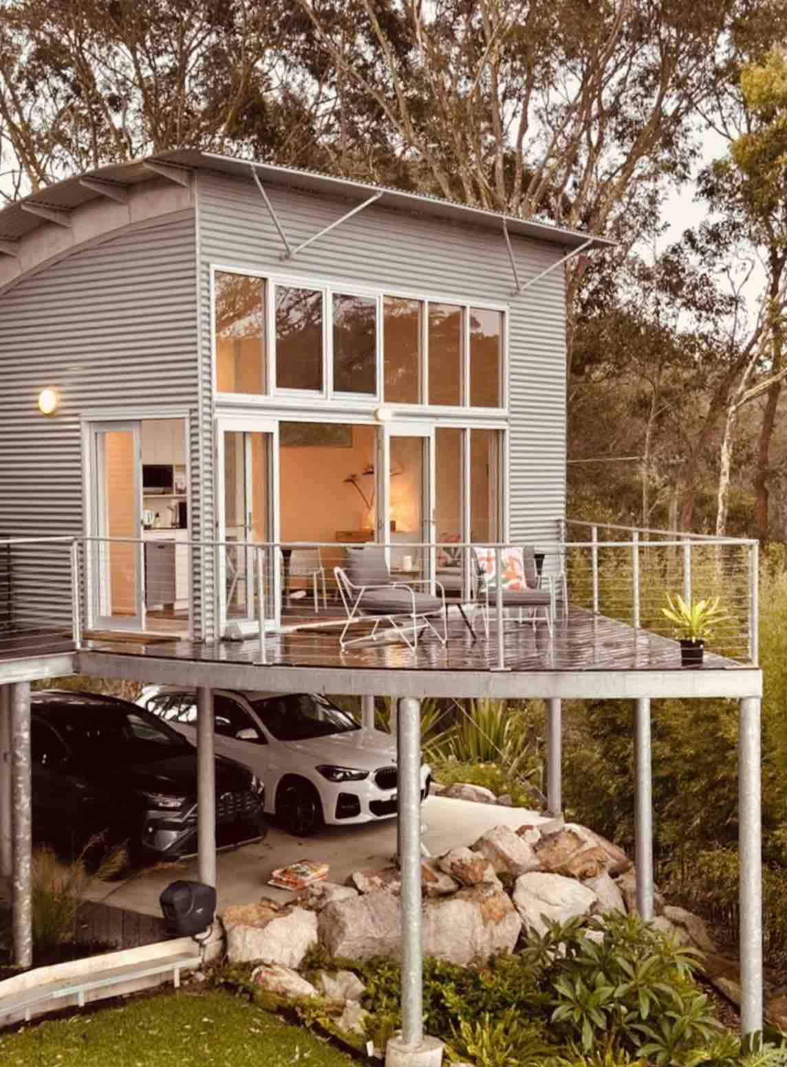 The unit is perched on stilts, featuring a spacious deck with outdoor seating. Large floor-to-ceiling windows offer a view of the inviting interior. A car park is located underneath, providing convenient access. Surrounding greenery adds to the peaceful ambiance of the location.