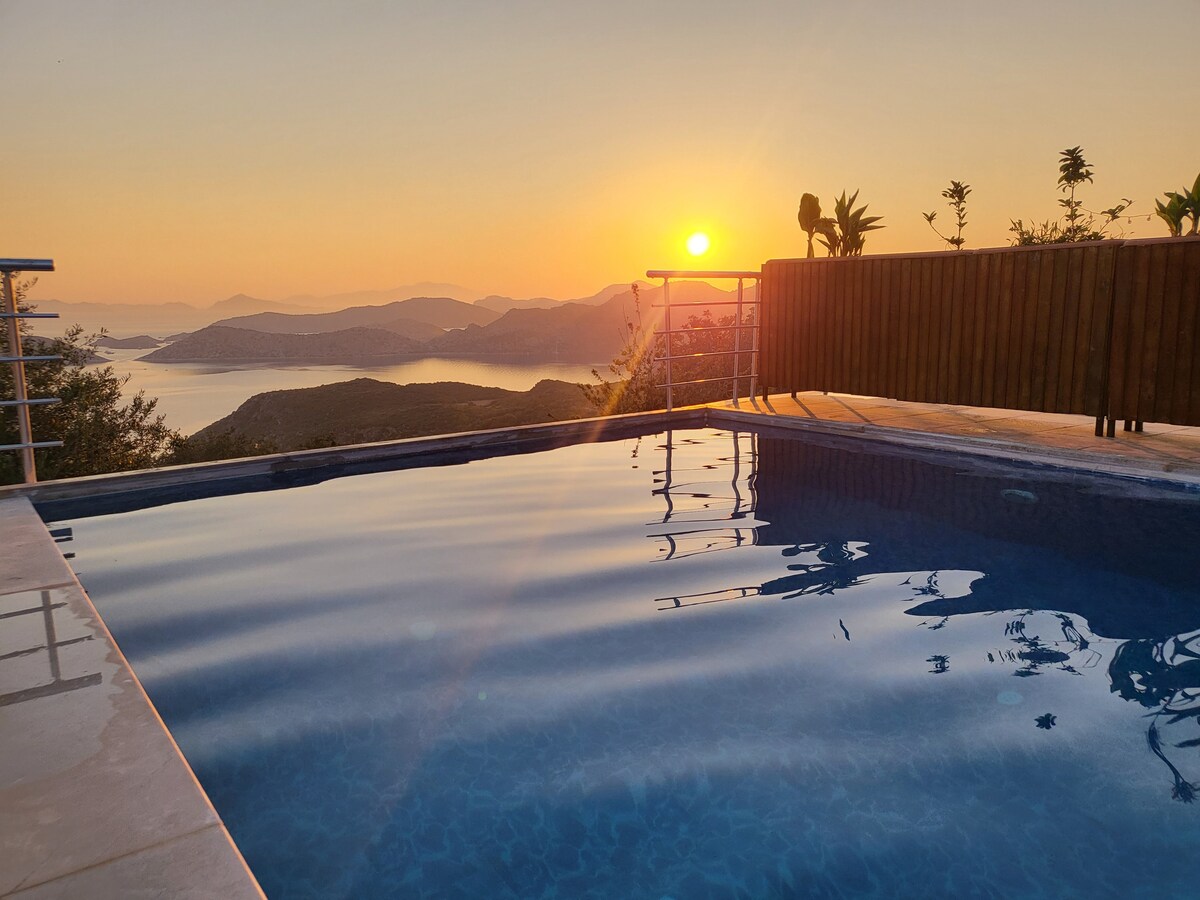 A serene view of a swimming pool reflects the warm hues of a sunset. The surrounding landscape features rolling hills and distant mountains, providing a tranquil backdrop. A wooden railing frames the pool area, enhancing the peaceful atmosphere.