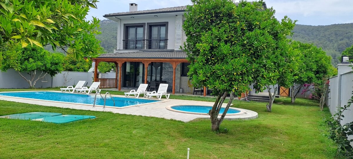 The exterior showcases a spacious lawn featuring a swimming pool and a children's pool. Lounge chairs surround the pools, and the house is framed by lush greenery. The upper level includes balconies with views of the surrounding area, adding to the outdoor experience.