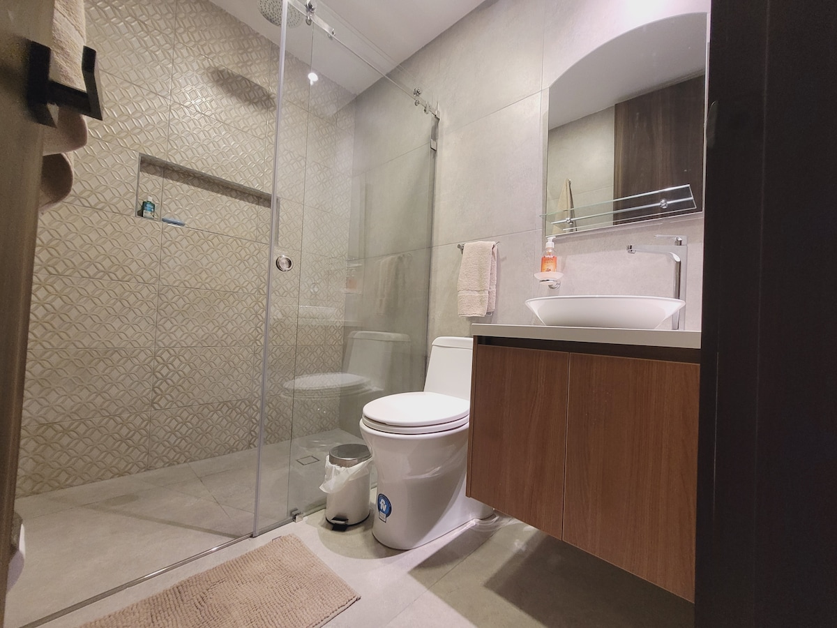 The bathroom features a glass-enclosed shower with textured tiles and a modern sink mounted on a wooden cabinet. A mirror is positioned above the sink, and a towel hangs on the wall nearby. Soft lighting contributes to a functional yet comfortable space.