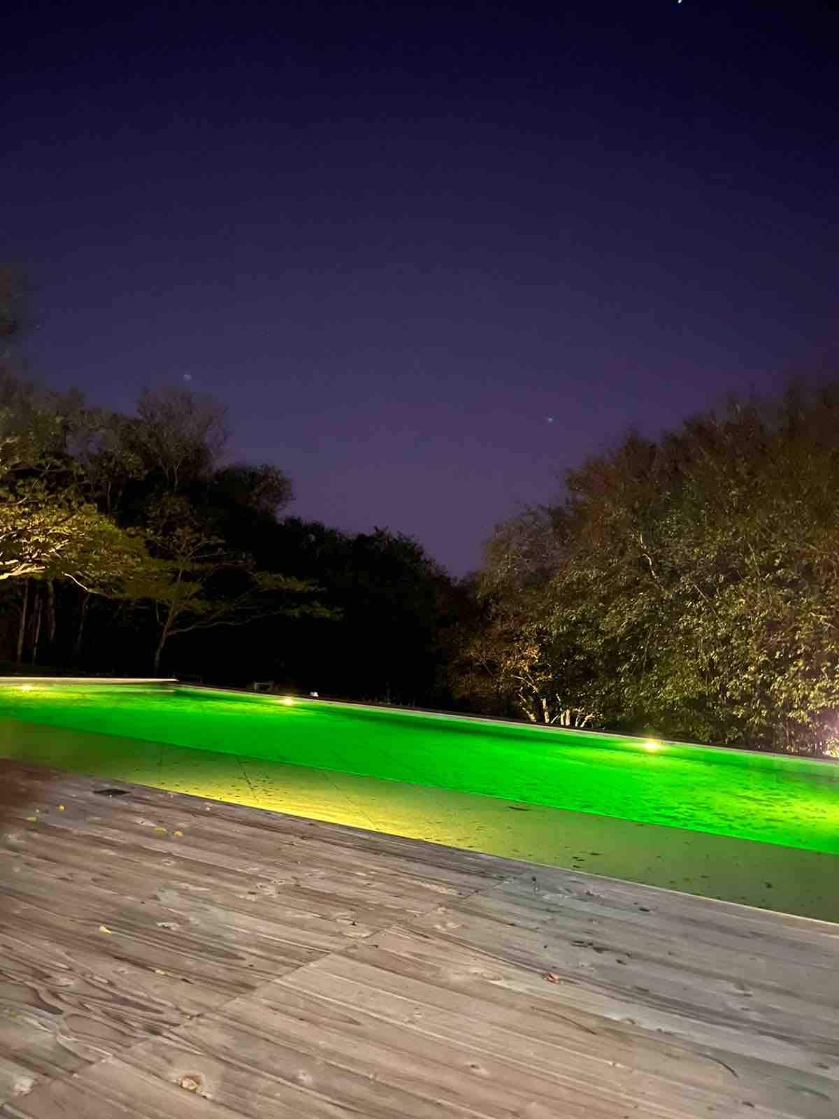 A tranquil view of an infinity pool, reflecting soft green lights against a serene backdrop of trees at dusk. The wooden deck surrounds the pool, providing a peaceful area for relaxation. The evening sky is deepening into twilight.