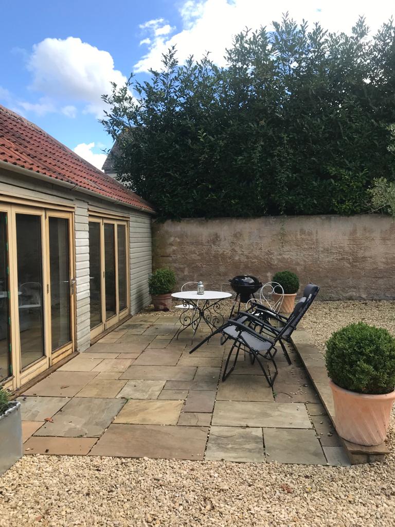 A courtyard area is presented with stone flooring, featuring a small round table and two chairs. Potted shrubs add greenery, while a barbecue grill is positioned nearby. The wooden structure with large glass doors reflects a welcoming outdoor space adjacent to the studio.