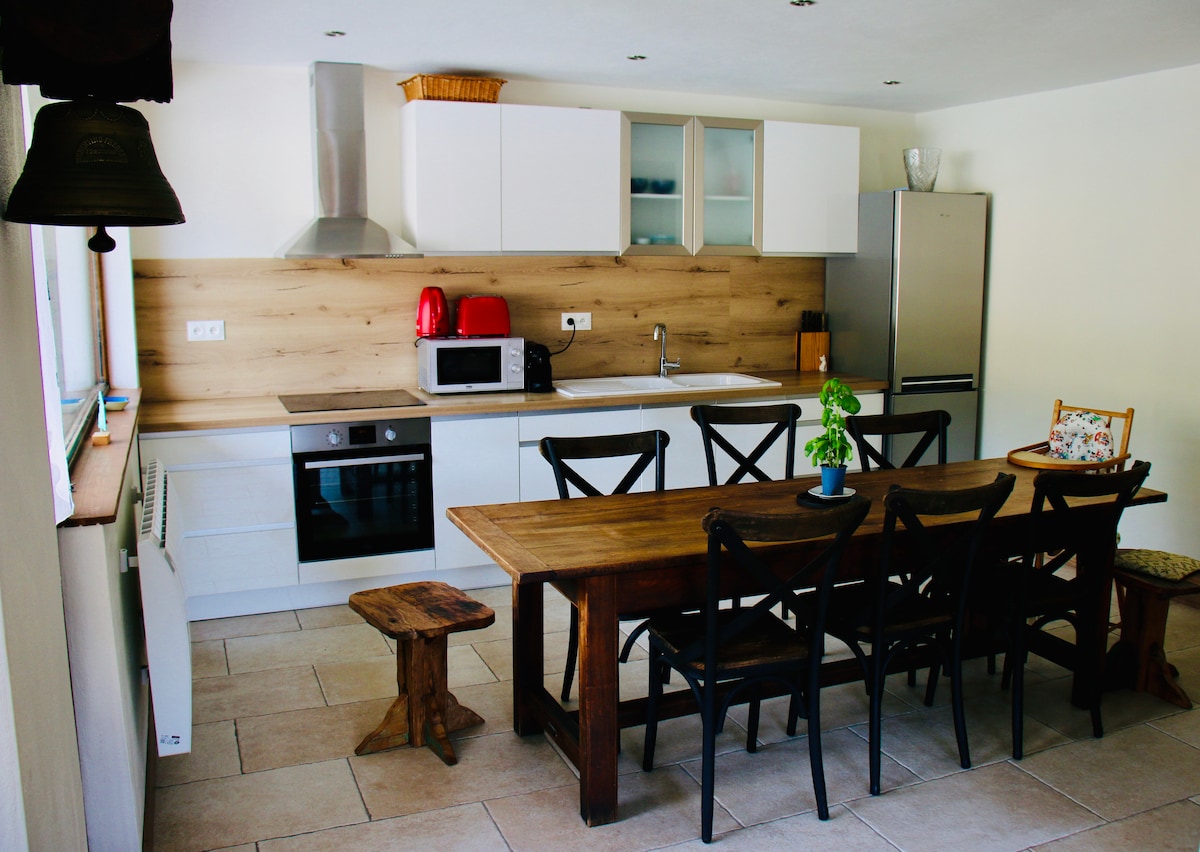A modern kitchen featuring a wooden dining table surrounded by black chairs. White cabinetry and a stainless steel fridge complement the warm wooden accents. Essential appliances, including a microwave and oven, are neatly arranged. Natural light enhances the space through a nearby window.