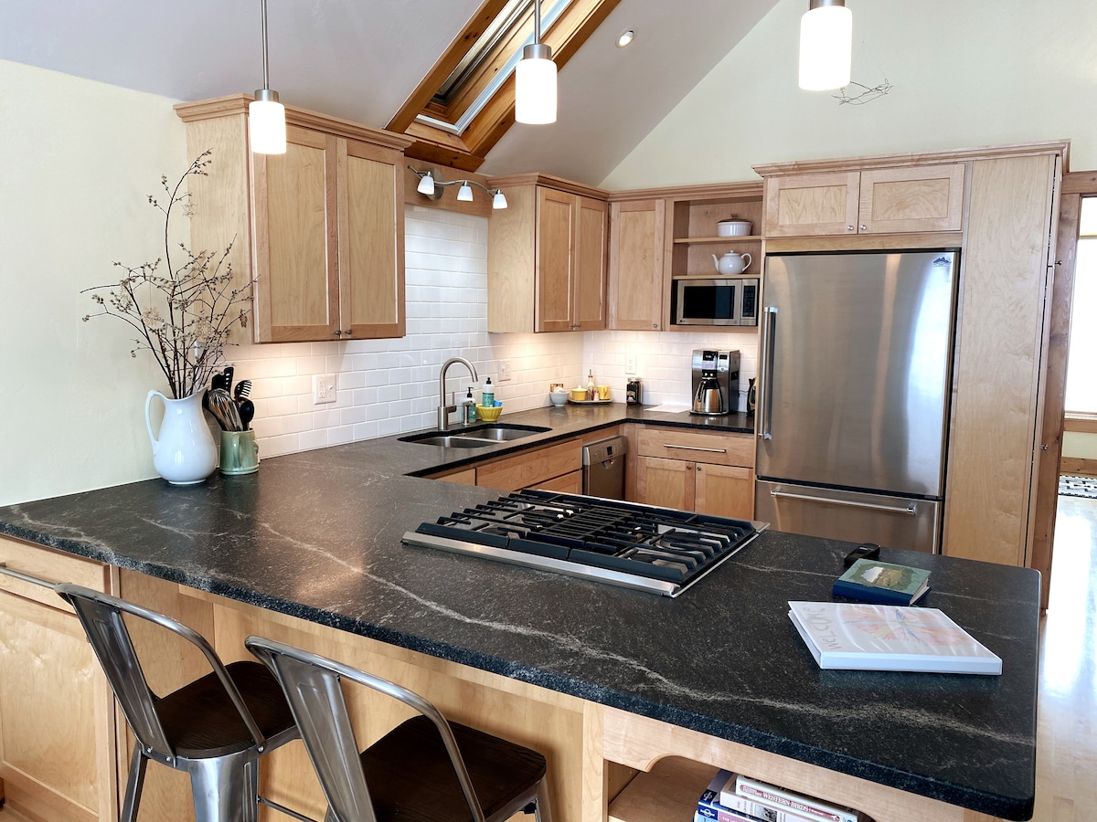 The kitchen features natural wood cabinetry and a spacious countertop with modern appliances. A gas stovetop is positioned on the island, accompanied by two metal bar stools. Light fixtures hang above, complementing the large window that allows for ample natural light.