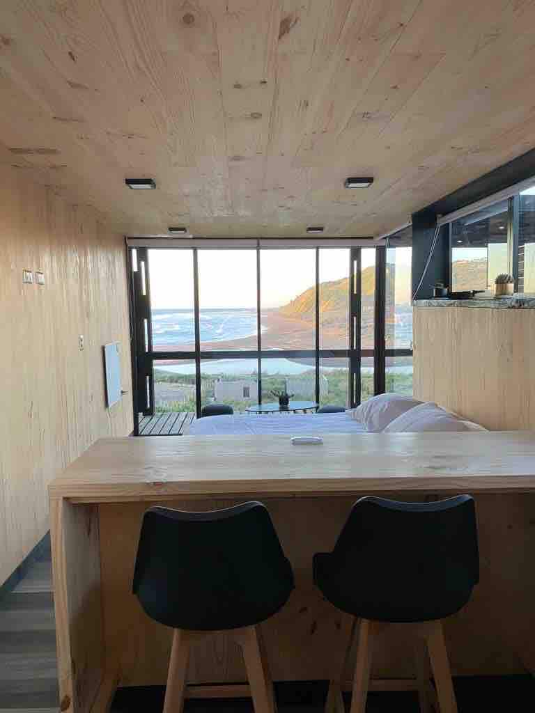 A modern interior features warm wooden walls and a panoramic window showcasing a stunning sea view. A small dining area with two sleek chairs is positioned near the window, complementing the open layout of the space, which includes a cozy sleeping area.