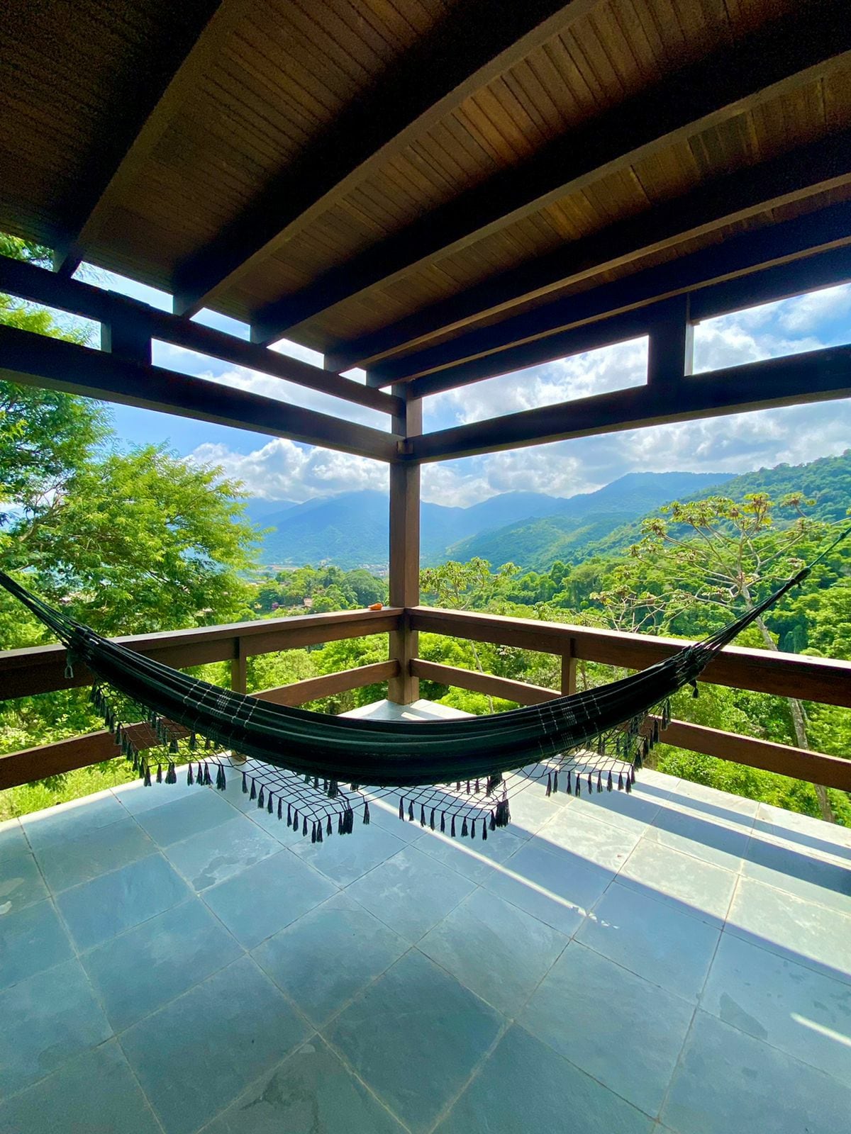 A shaded balcony is framed by wooden beams, featuring a hammock suspended between supports. The view showcases lush mountains and greenery under a bright sky with scattered clouds, creating a serene outdoor space for relaxation.