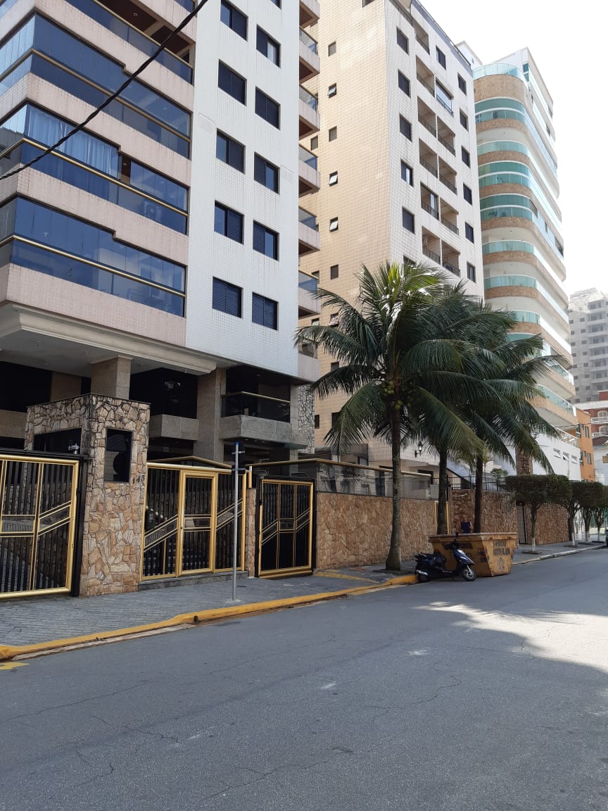 The image displays a multi-story residential building featuring a modern facade with large windows. Palm trees are visible near the entrance, which is framed by decorative stonework and gates. The street is lined with a mix of buildings, indicating an urban environment.