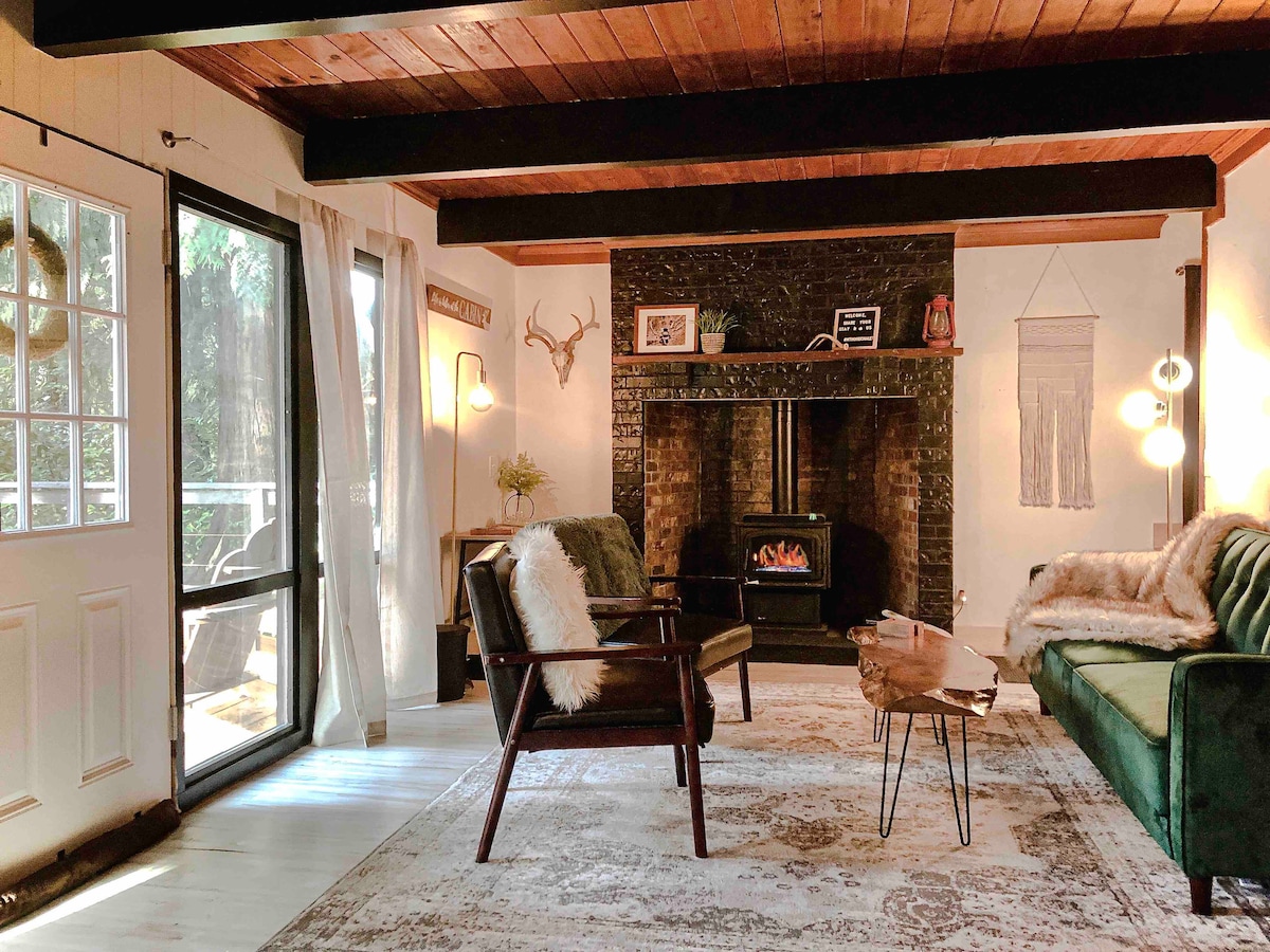 A cozy living area features vaulted ceilings and wood beams, with a propane fireplace highlighted against a dark brick wall. Comfortable seating is provided by an armchair and a plush sofa, arranged on a large area rug. Natural light comes through a door leading to the outdoor space.