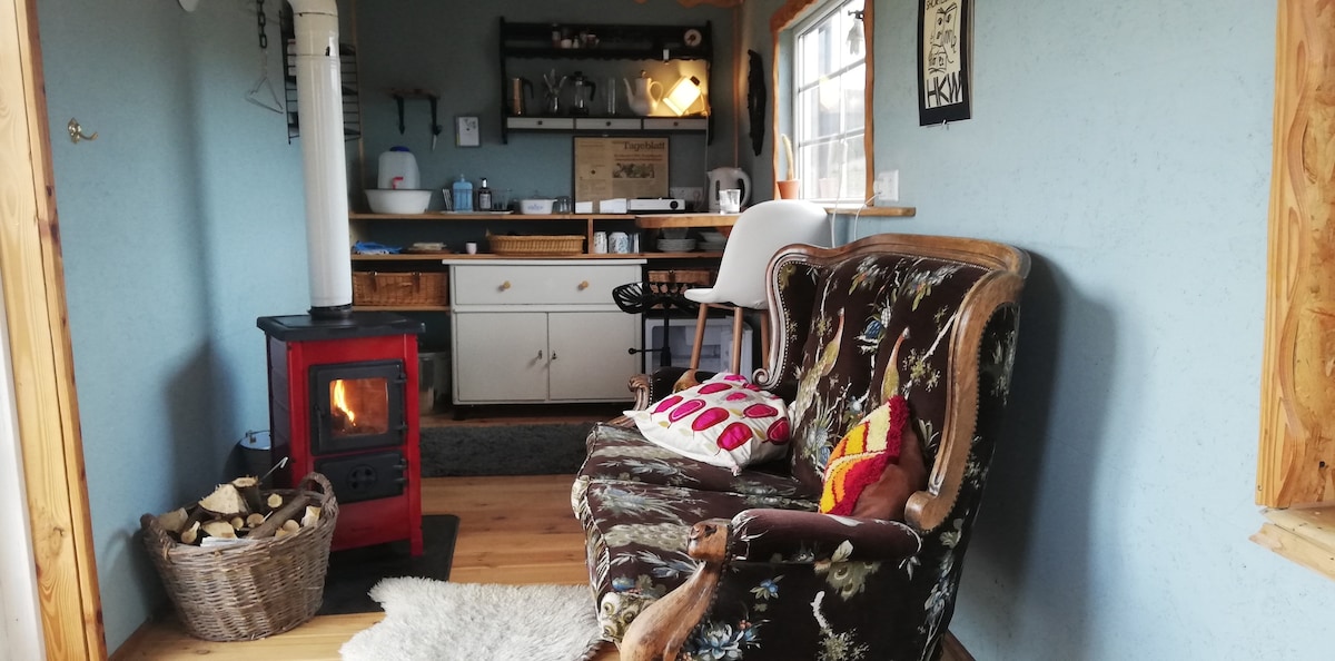 A cozy interior space features a vintage armchair adorned with colorful cushions. A small red stove provides warmth, complemented by a stack of wood nearby. The kitchen area includes shelves stocked with supplies, while natural light enters through a window, enhancing the tranquil setting.