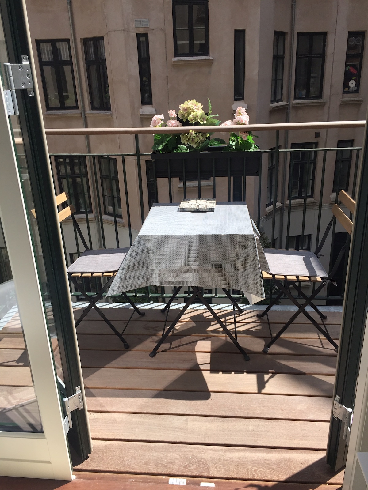 A balcony is depicted with a small, round table at its center, surrounded by two folding chairs. A light grey tablecloth covers the table, which is adorned with a decorative item. Green plants are visible in a planter, enhancing the calming outdoor space.