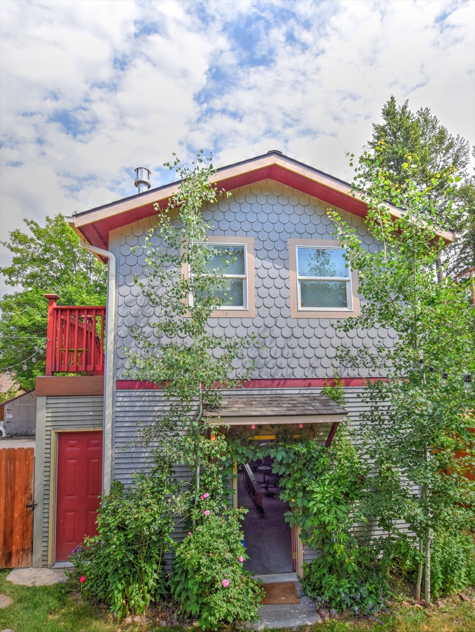 The exterior of the cabin features a unique patterned façade, complemented by two windows and a red door. Surrounding greenery includes trees and flowering plants, while a small staircase leads to a deck above, providing a welcoming entrance to the property.