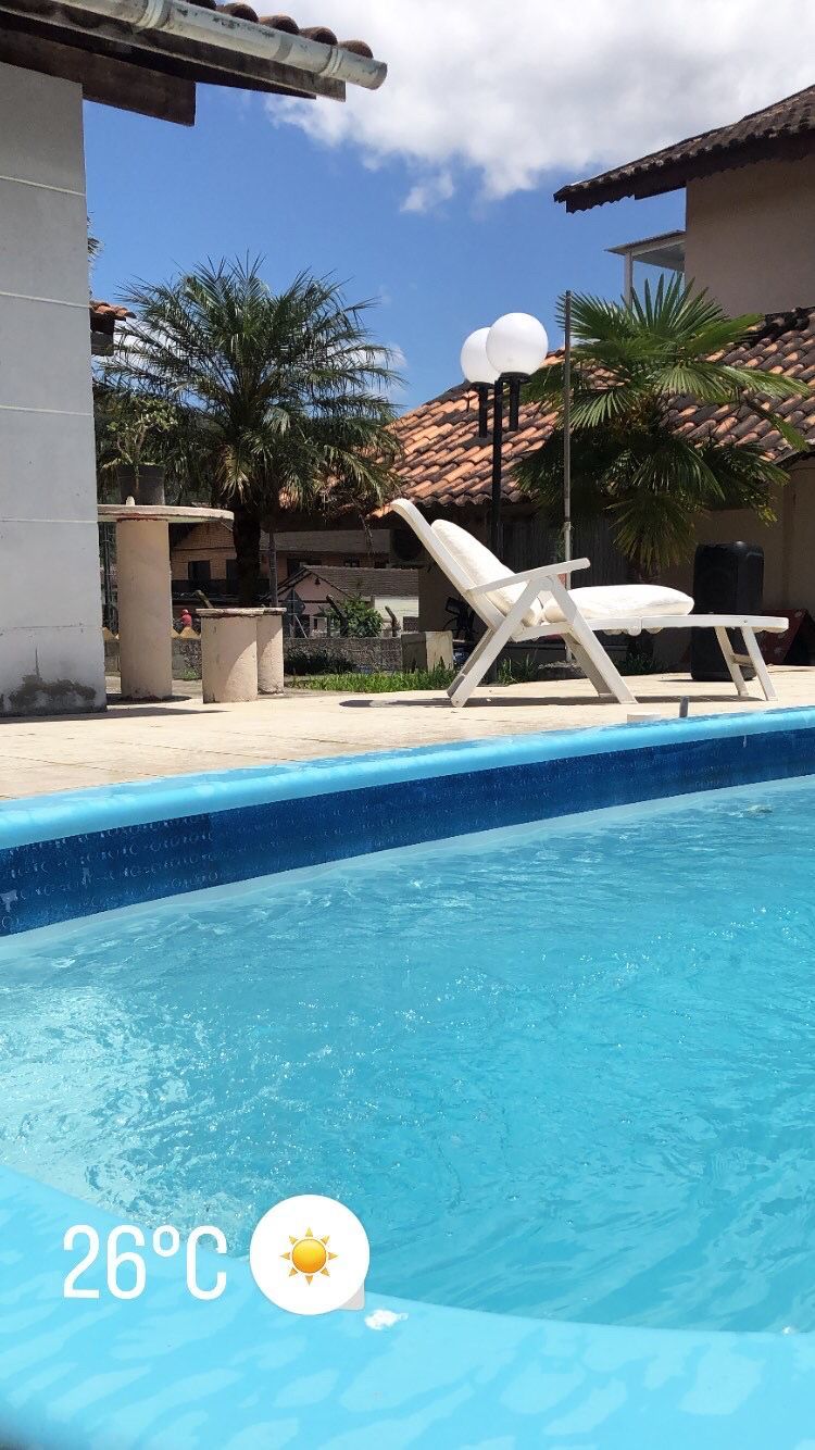 A clear blue swimming pool is visible in the foreground, with a white lounge chair positioned nearby. Lush greenery and tropical plants surround the area, while a partly cloudy sky adds a touch of brightness to the scene.