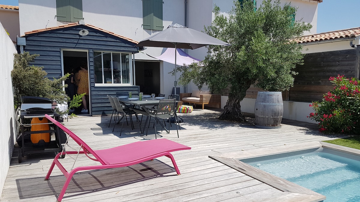 An outdoor terrace features a wooden deck surrounding a private pool. A pink lounge chair rests beside the pool, and a dining area is set with a large table and chairs. Shade is provided by a large umbrella, while greenery borders the space.