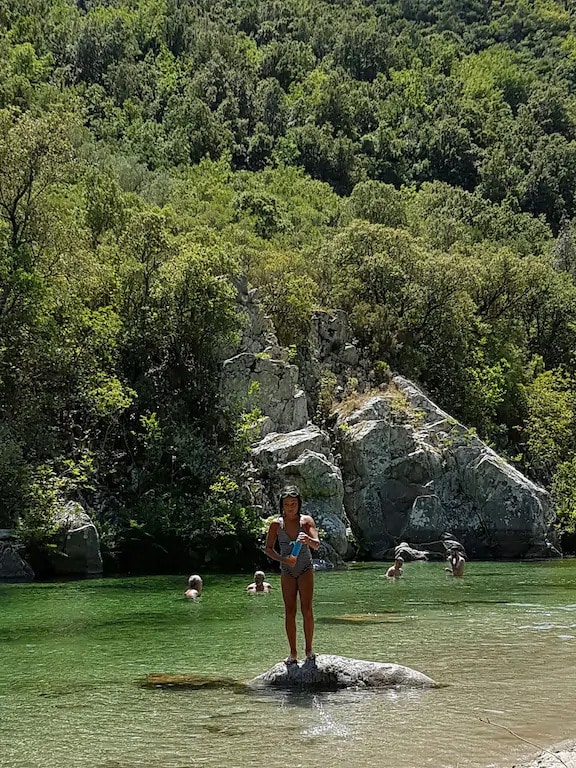 A woman stands on a rock in a serene river, surrounded by lush greenery and rocky formations. The crystal-clear water reflects the vibrant hues of the landscape. In the background, individuals are seen enjoying the water, highlighting a peaceful natural setting.