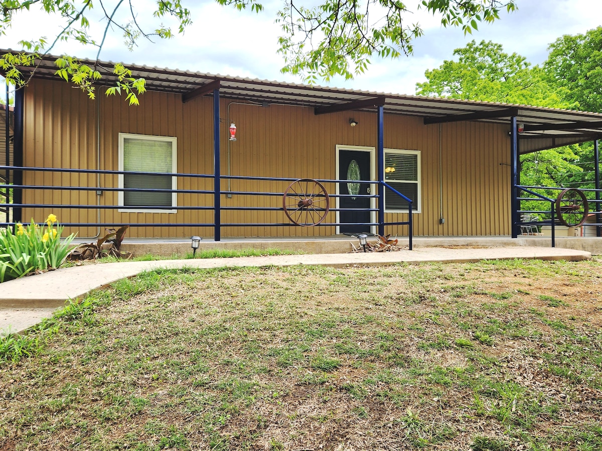 The exterior of a barndominium is visible, featuring a rustic design with a beige facade and dark roof. A covered porch is framed by decorative wheels and outdoor seating, set against a backdrop of lush greenery and a well-maintained lawn.