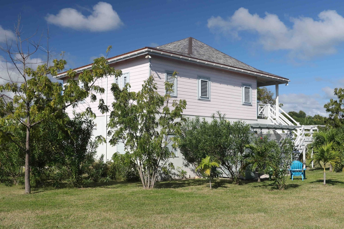 The exterior of the apartment is showcased, featuring a two-story structure with pink siding and white trim. Lush green landscaping surrounds the building, with several trees and shrubs. A blue chair is positioned in the yard, complementing the peaceful setting under a partly cloudy sky.