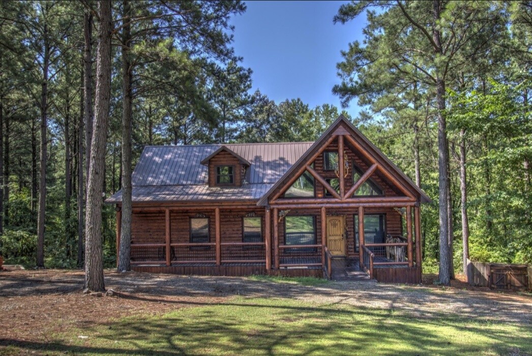 The cabin is set amongst tall pine trees, exhibiting a rustic wooden exterior with large windows and a covered porch. Its triangular roof and architectural details reflect a log cabin style, while the surrounding greenery enhances the natural setting.