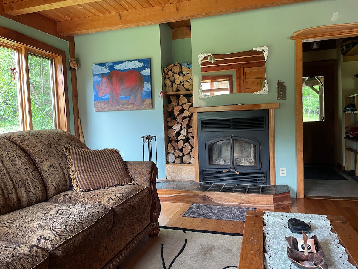 A comfortable living area is featured with a plush sofa adorned with patterned cushions. A fireplace is visible, flanked by neatly stacked firewood. A piece of artwork depicting a red cow decorates the wall, and natural light streams in through large windows, illuminating the wooden interior.