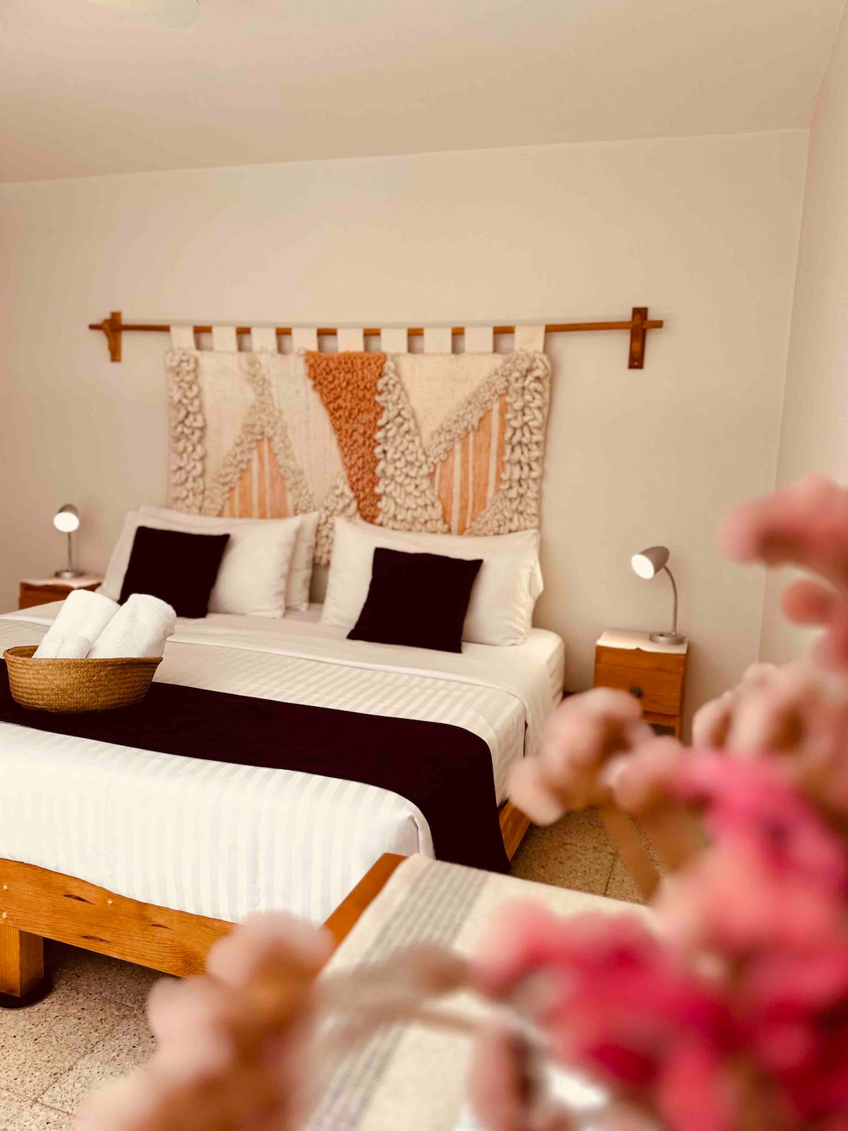A comfortable bedroom features a spacious bed adorned with crisp linens and decorative pillows. A textured wall hanging adds a touch of warmth. Two bedside lamps provide soft lighting, and a woven basket filled with fresh towels is positioned on the wooden nightstand.