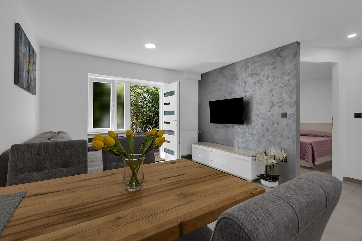 A dining area features a wooden table surrounded by upholstered chairs. White walls are complemented by a grey accent wall, and a flat-screen TV is mounted for entertainment. A window invites natural light and offers views of the garden, while the entrance to the bedroom is visible.